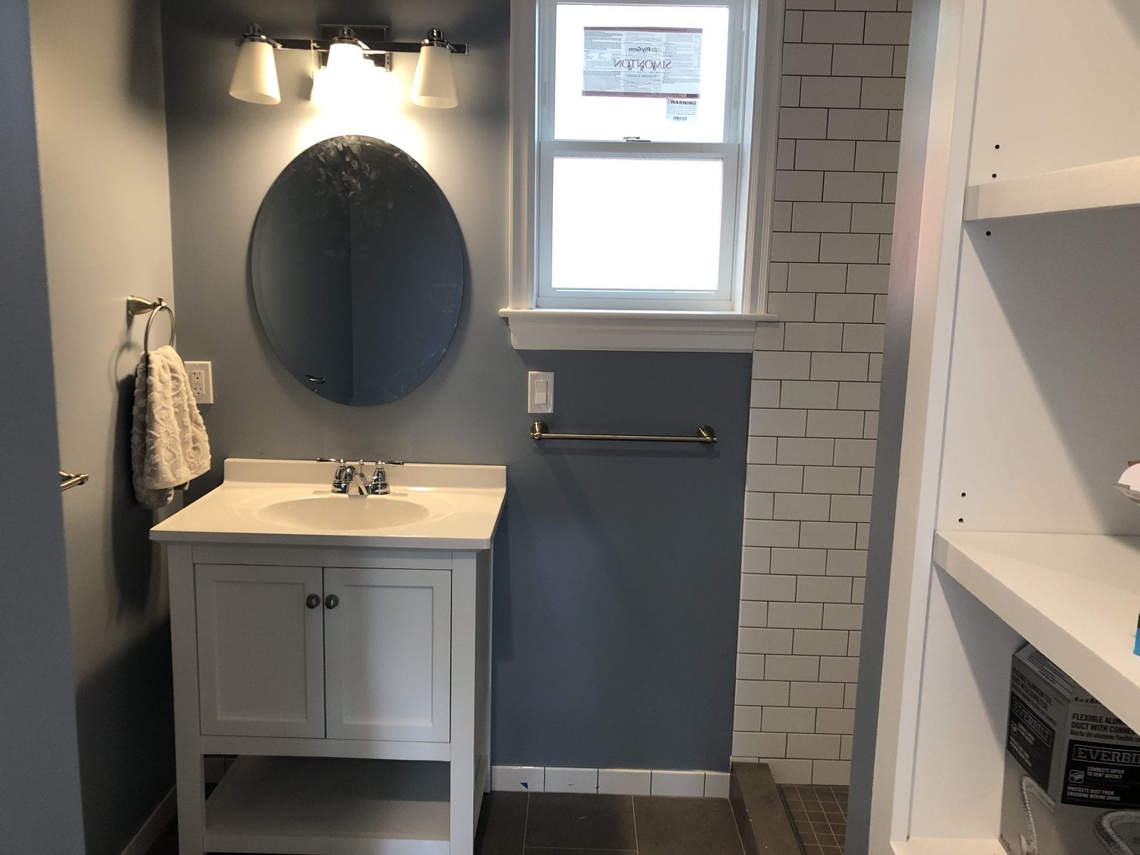 Completed bathroom with a white shaker vanity, oval mirror, three-light fixture and a white subway tile shower surround against slate-blue walls