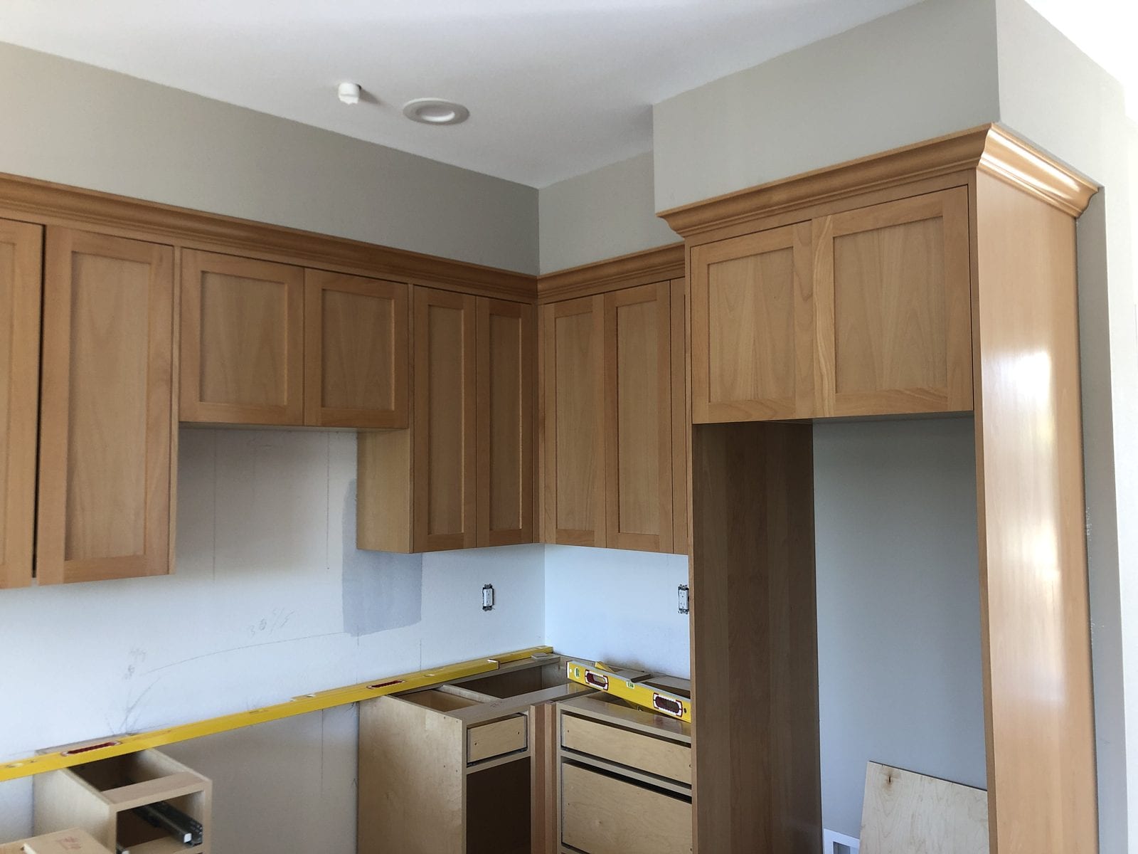 Maple shaker wall cabinets and crown moulding installed in a kitchen, with base cabinets set and countertops not yet fitted