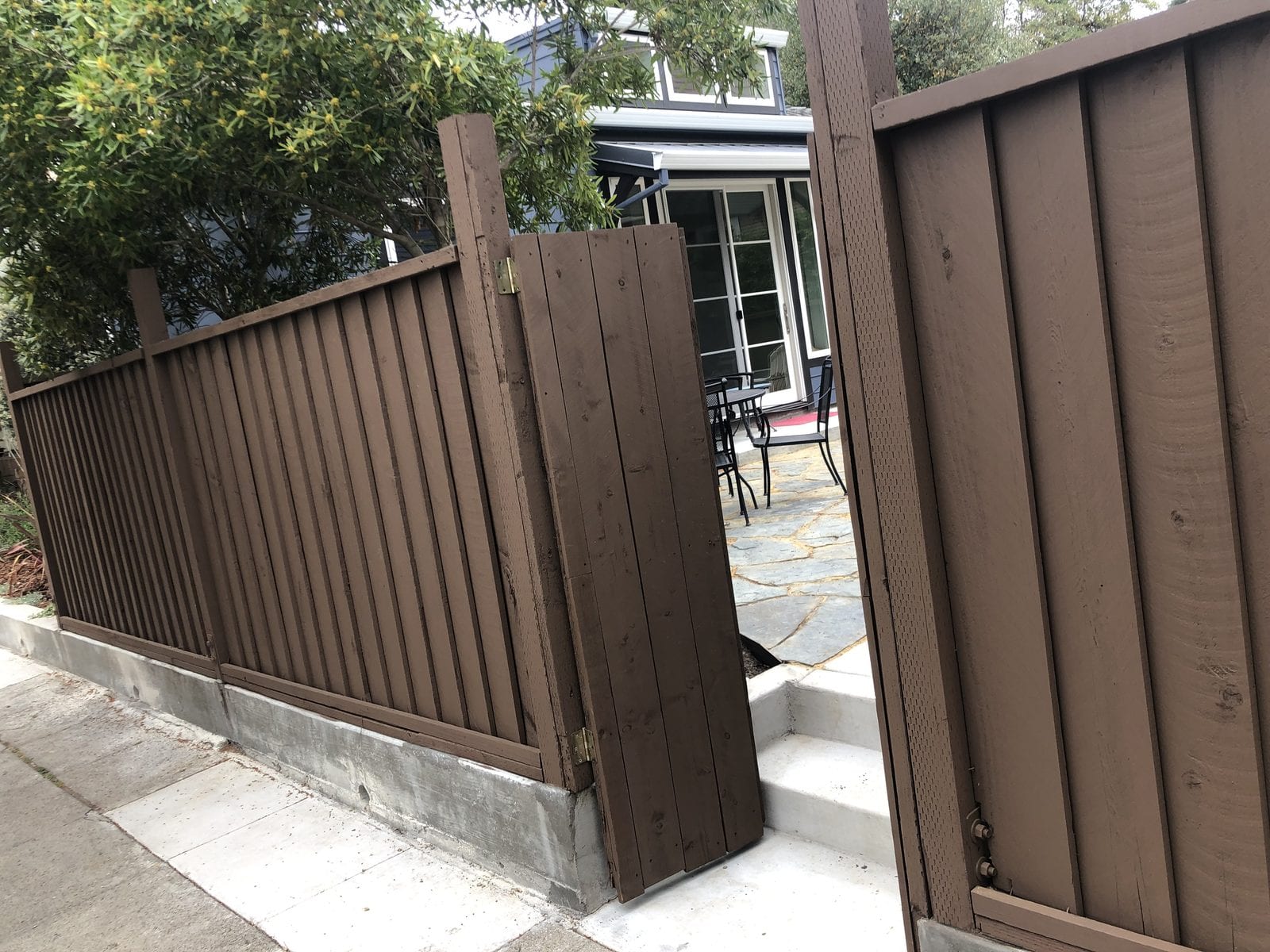 Board-on-board timber fence and matching gate stained dark brown on a poured concrete curb, opening onto a flagstone patio