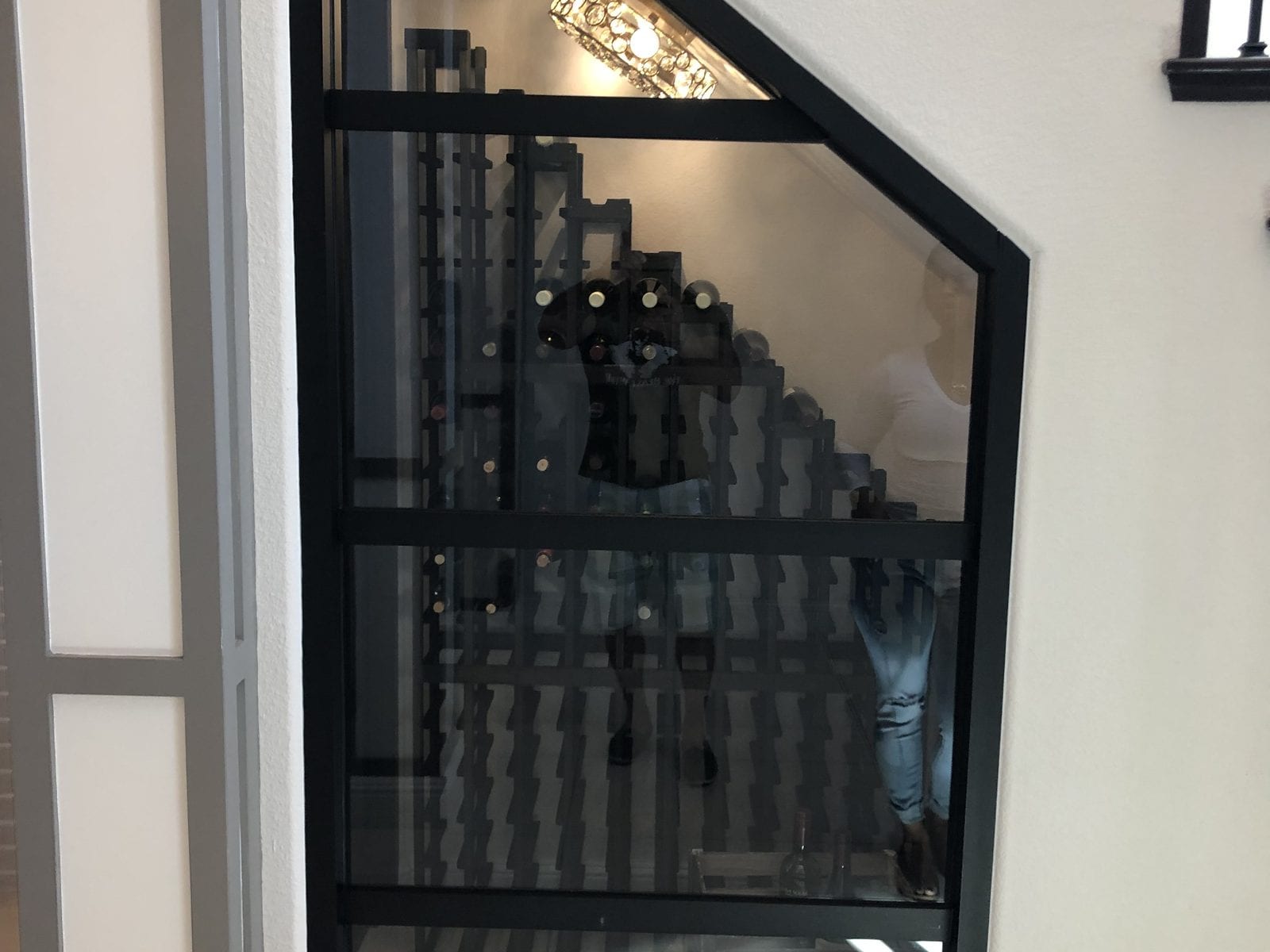 Climate-glazed wine store built into the space under a staircase, with a black steel-framed glass door, interior lighting and tiered wooden bottle racking